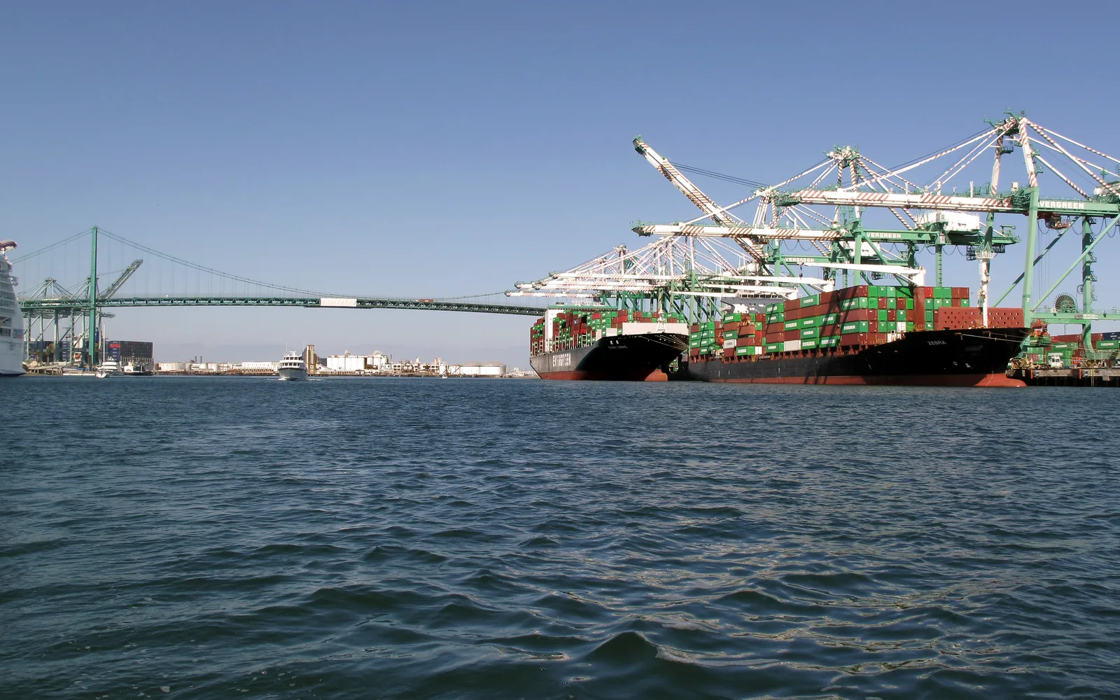 Container ships at loading cranes in the Port of Los Angeles, with the Vincent Thomas Bridge visible across the harbor